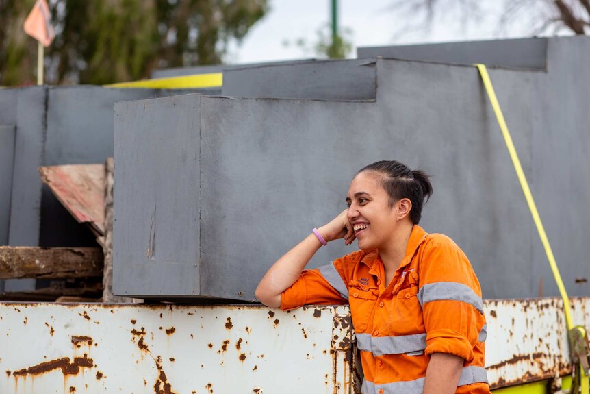 A woman in a high-visibility work shirt leans on large silver pillars loaded onto a trailer.