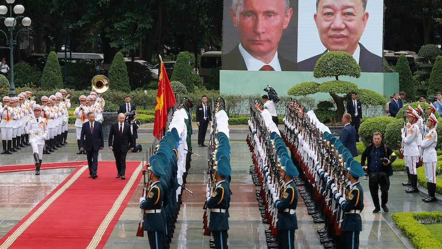 Two men walk along a red carpet while a brass band plays behind them and men in military uniform watch on.