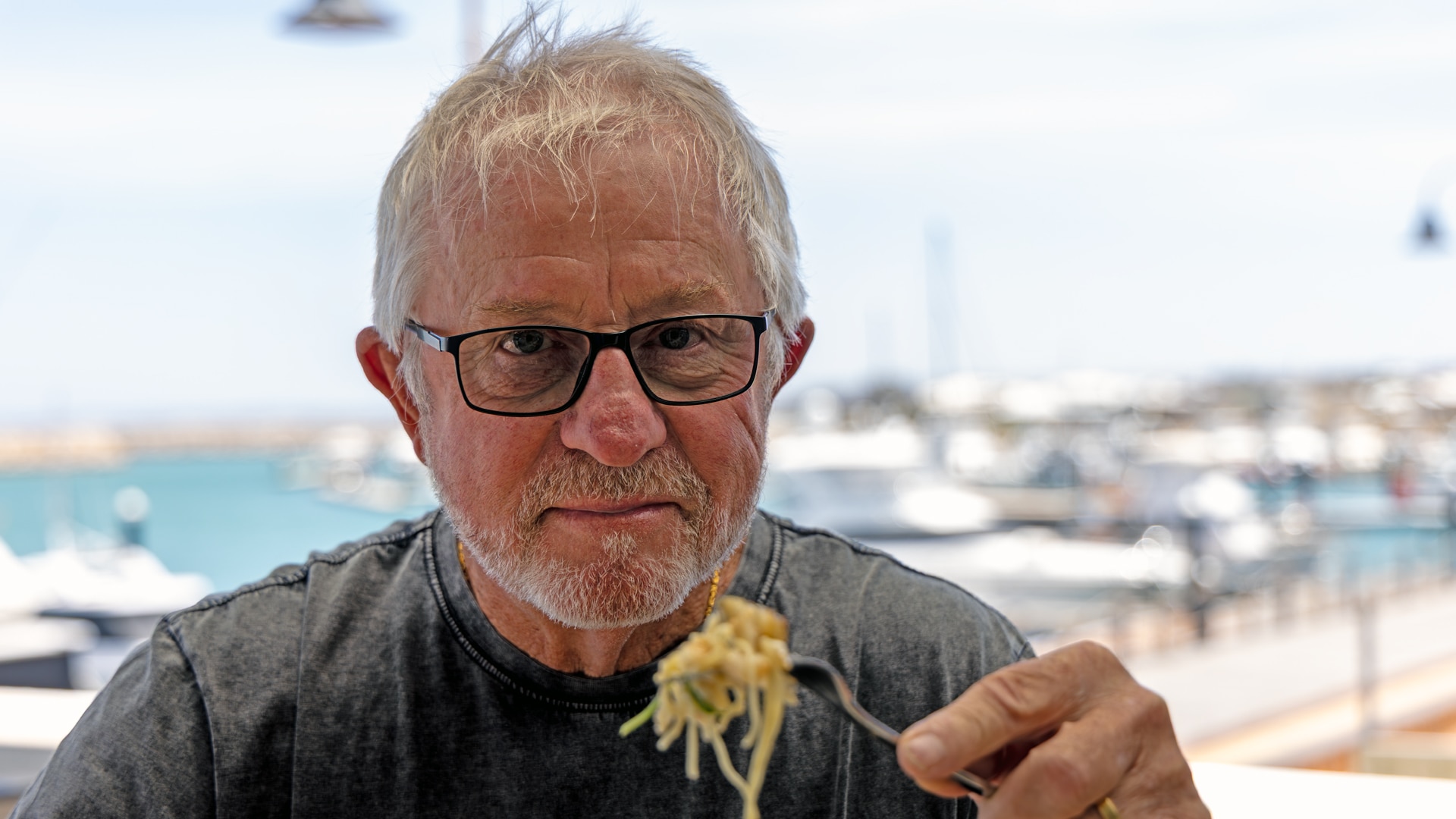 Un hombre con gafas y pelo corto y fino mira a la cámara. Sujeta la pasta y el pescado con un tenedor. 
