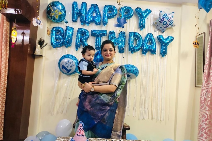 A woman wearing a sari holds a boy in front of a sign that says Happy Birthday.