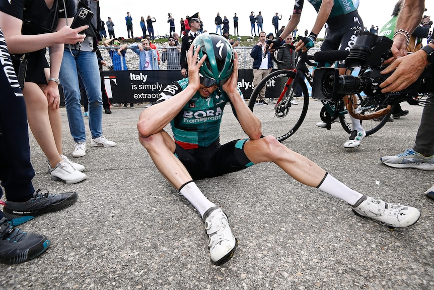 Jai Hindley sostiene su cabeza con casco mientras se sienta en el camino con las piernas abiertas frente a él.