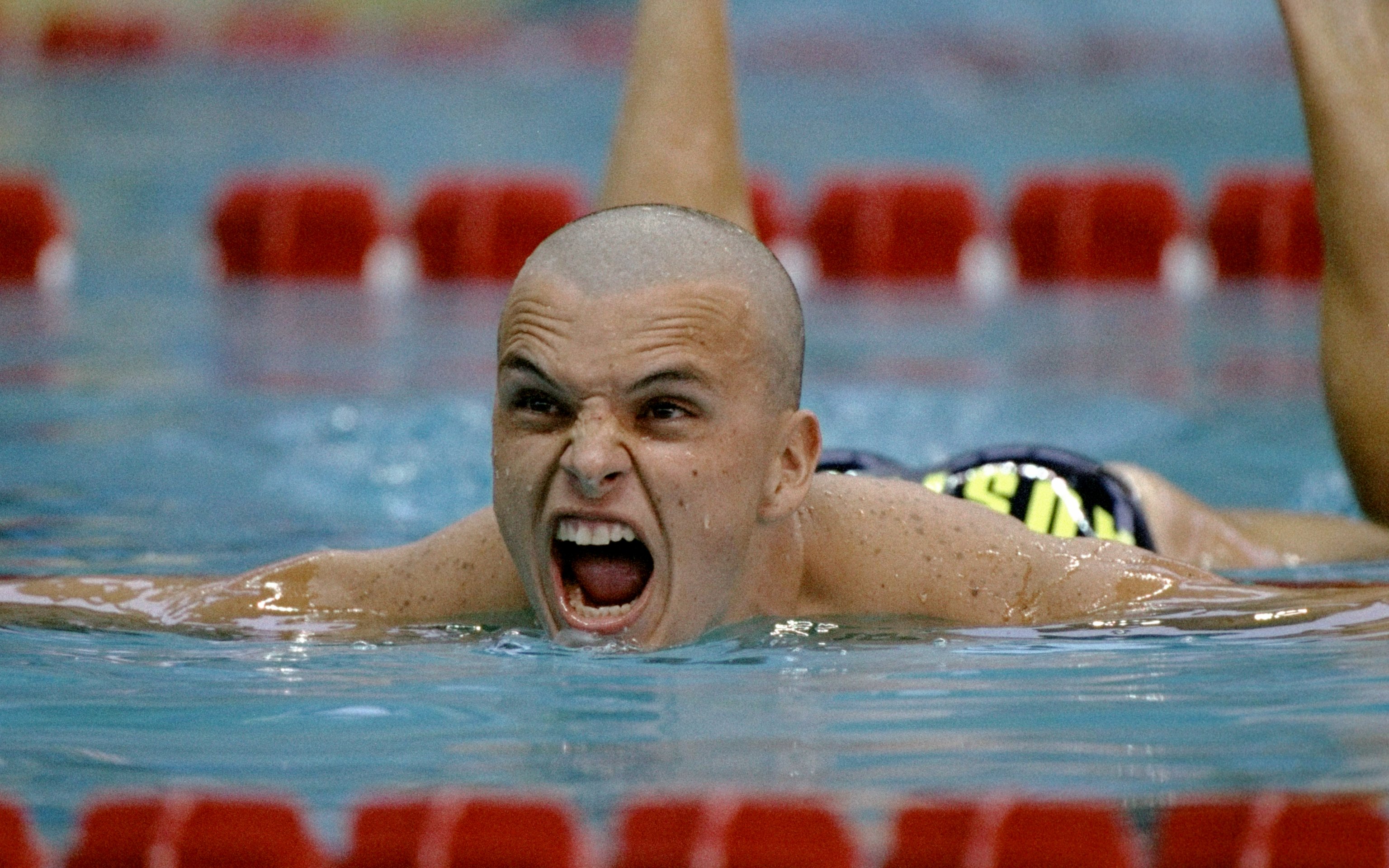 A bald man in a swimming pool yells while swimming over the red lane ropes.