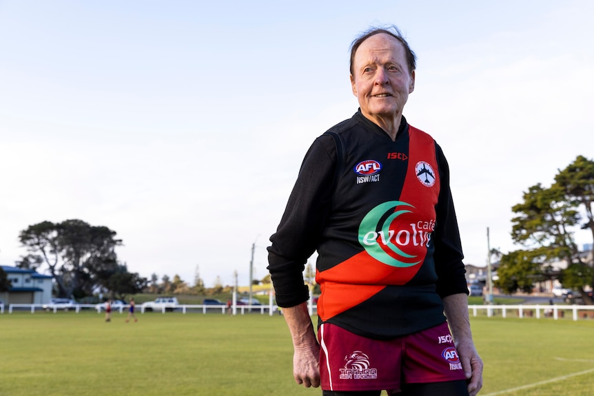 A man in a black and red Aussie Rules jumper stands in front of a footy oval