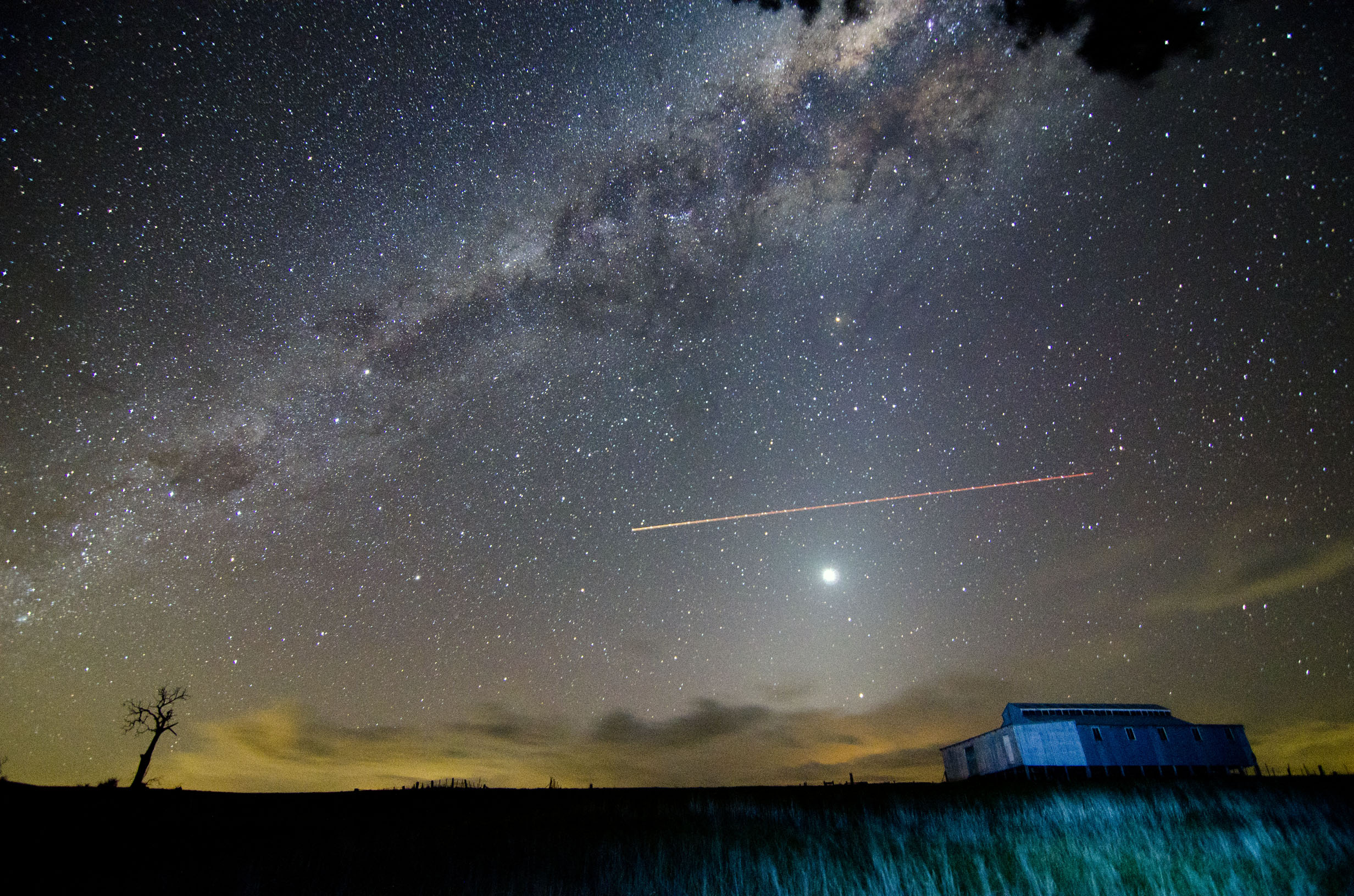 A bright light can be seen in a very starry night sky with a shearing shed in the foreground. 