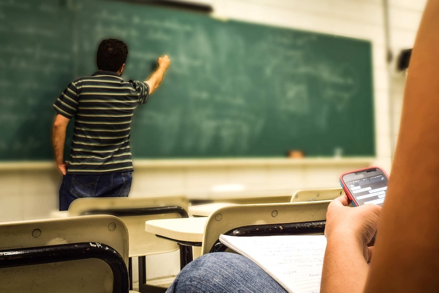 A man writes on a blackboard while a student sits in a chair looking at their mobile phone.