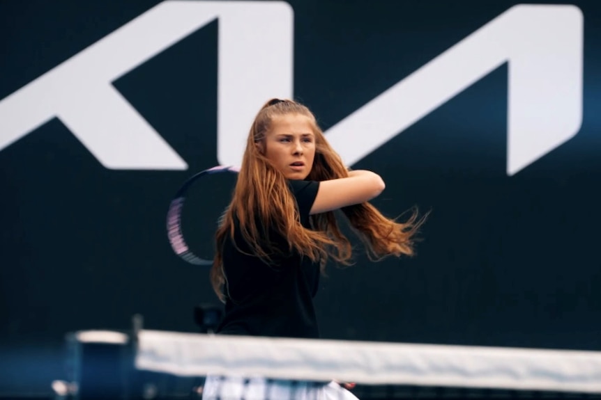 A young woman with a vision impairment preparing to hit a ball on a tennis court, long red hair half tied back.