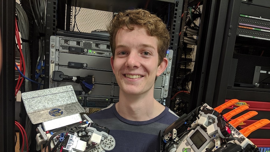 Young man stands in front of a electronics holding the robots he and his team designed