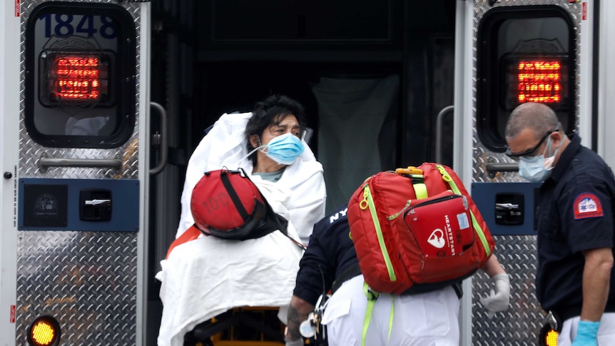 A woman with black hair sitting on a stretcher wearing a mask being loaded into an ambulance by two paramedics.
