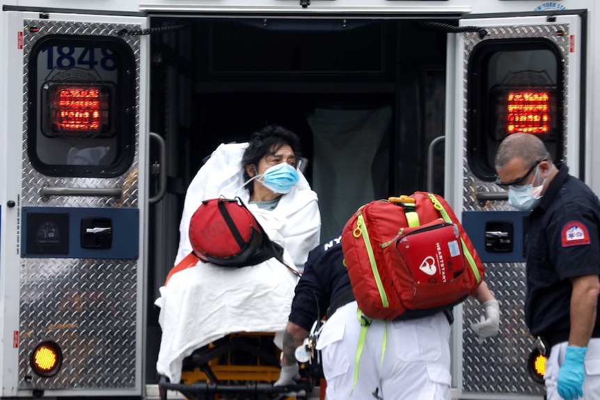 A woman with black hair sitting on a stretcher wearing a mask being loaded into an ambulance by two paramedics.