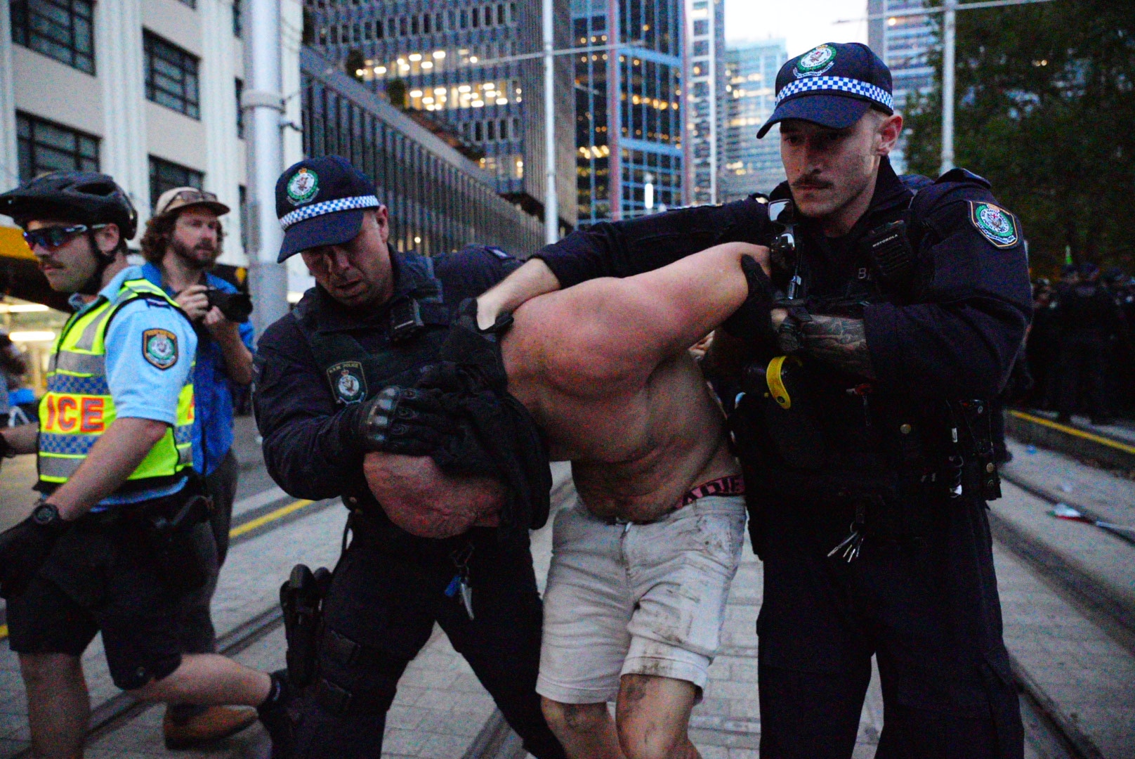 Dos agentes de policía retienen a un hombre sin camiseta y con pantalones cortos sucios.