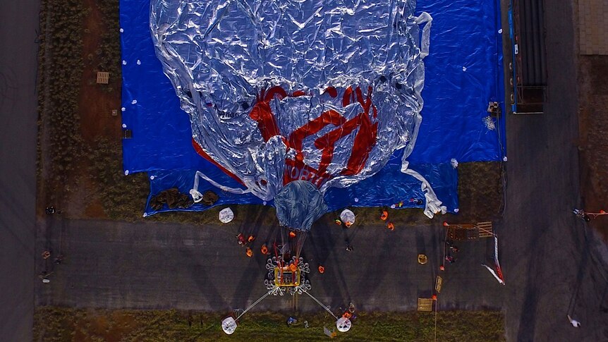An aerial picture of a deflated silver hot air balloon on the ground on a blue tarpaulin.