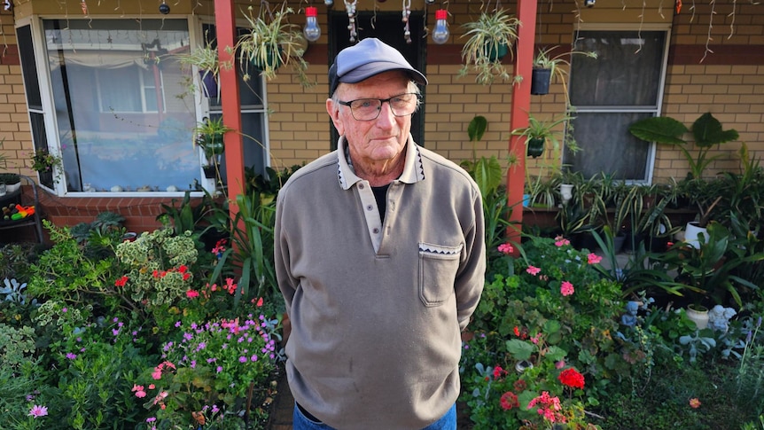 A mean wearing a grey sweater, glasses, and baseball cap standing in front of a brick house with garden