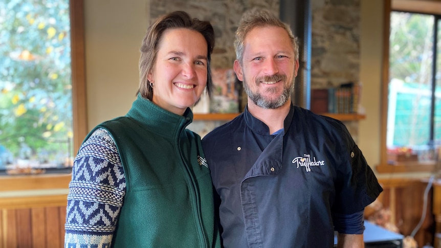 A woman in a green vest standing next to a man in a black shirt in a restaurant with a fire place. 