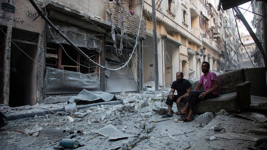 Two men sit on an old couch looking at the rubble around them.