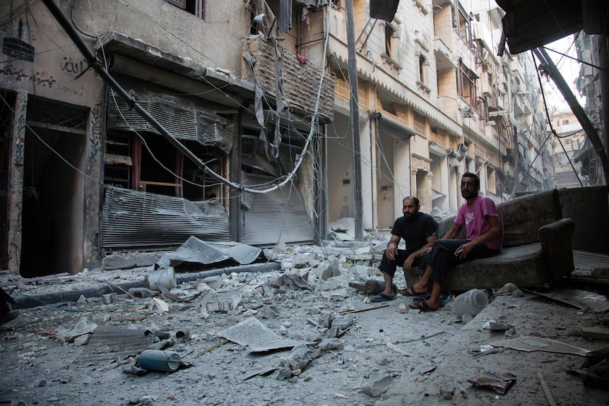 Two men sit on an old couch looking at the rubble around them.