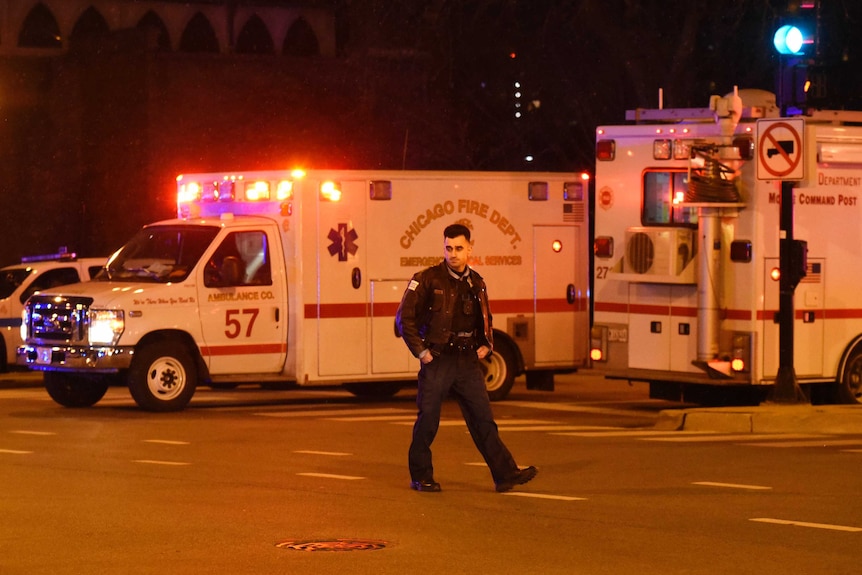 A lone police officer walks by a line of emergency vehicles with a solemn expression on his face.