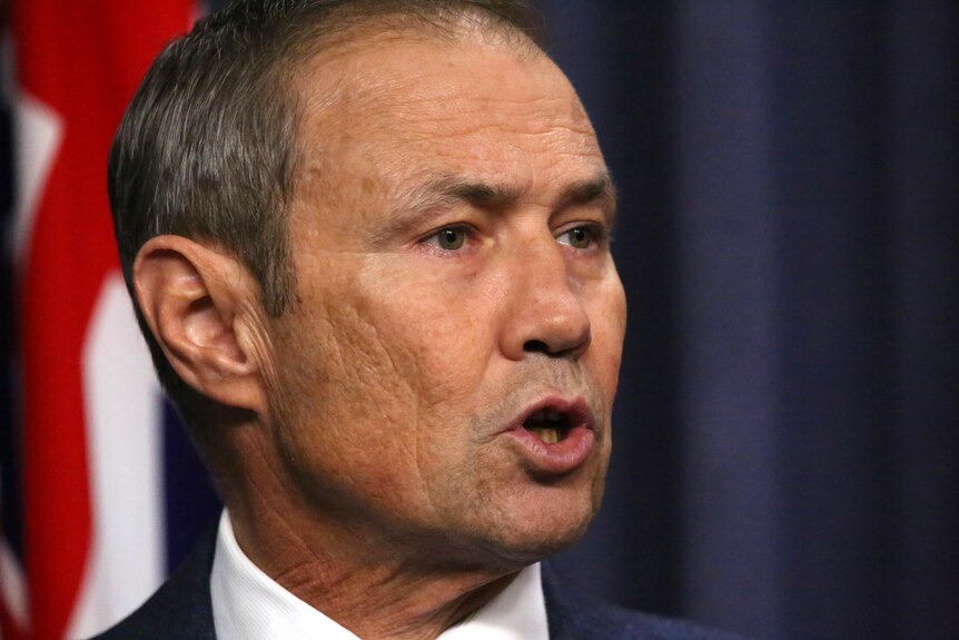 A tight head shot of Roger Cook speaking while looking to his left, with a union jack on a flag behind him.