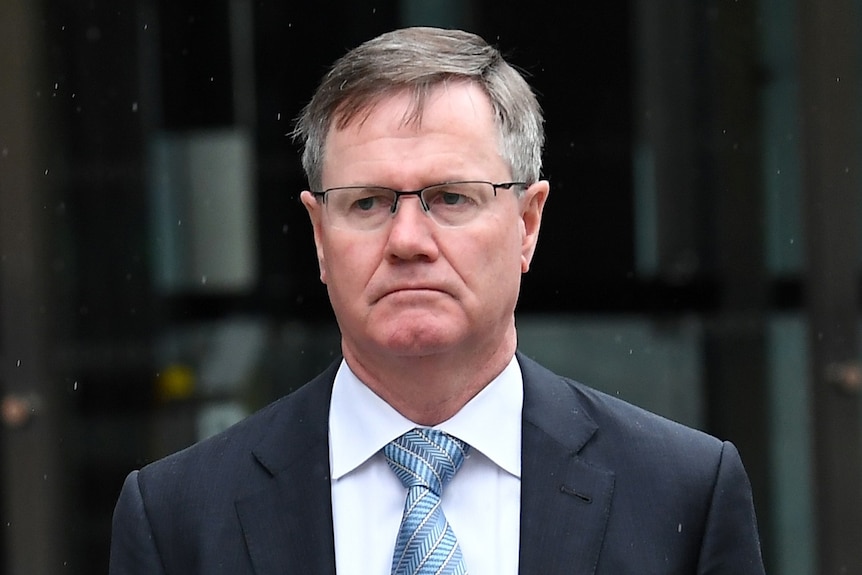 Stephen Healy is dressed in a blue tie and suit as he walks out of a court building under light rain.
