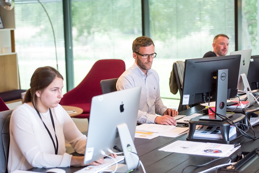 A photo shows three workers at their computers, sitting along a shared desk