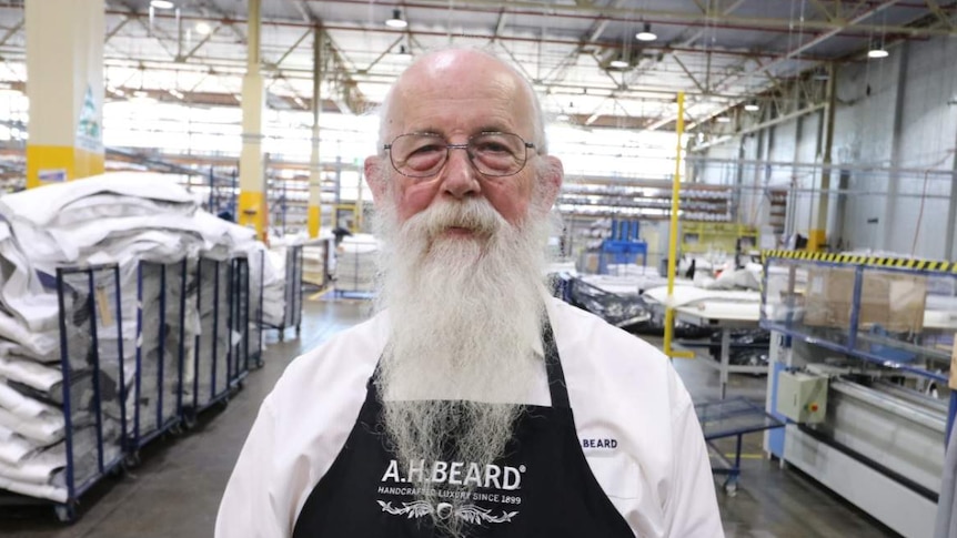 A man with a long white beard, wearing an apron with the A.H. Beard logo, standing in a mattress factory.