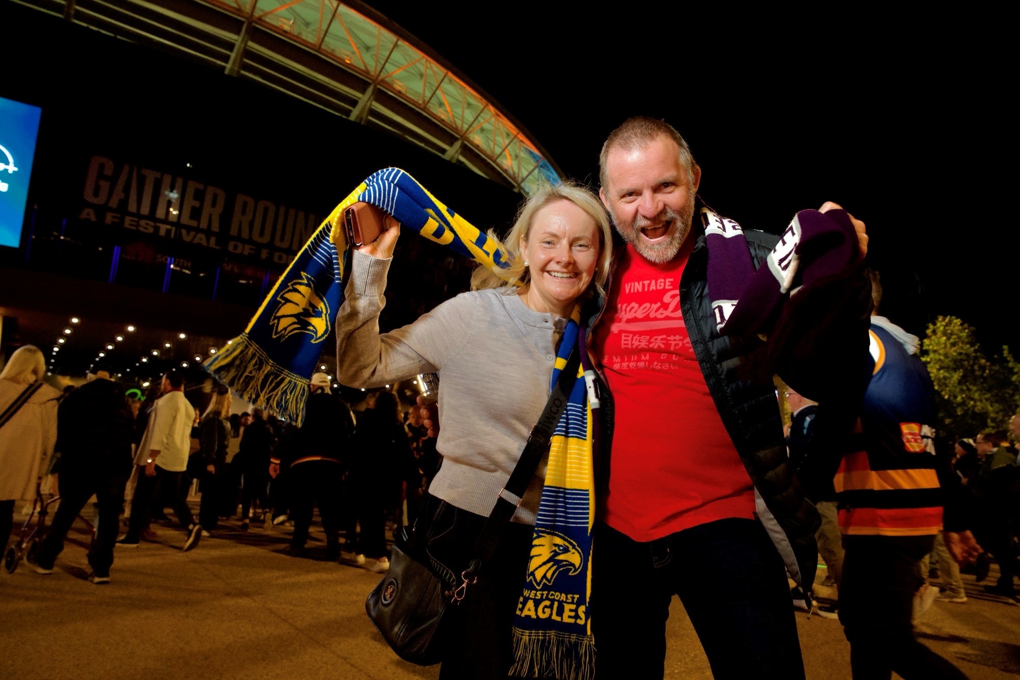 Uma mulher loira e um homem careca estão abraçados, segurando seus lenços de futebol, torcendo. 