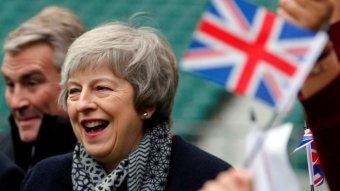 Theresa May is seen laughing while a person waves a small British flag that is out of focus