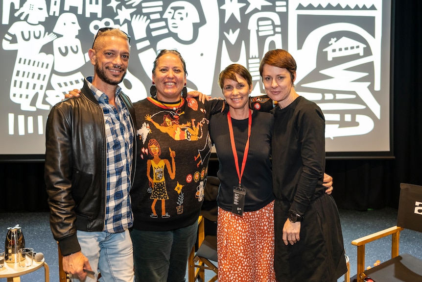 Four people, arms around each other, standing on stage smiling at the camera, the Birrarangga artwork behind them.