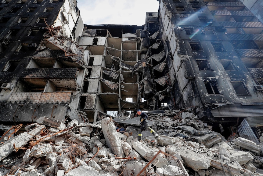 A person walking over rubble in front of building damaged by war.