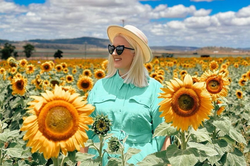 A woman with blong hair, a green dress and love heart sunglasses stands in a field of big yellow sunflowers.