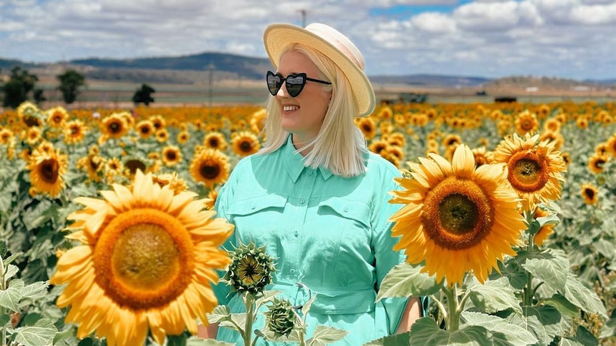 A woman with blong hair, a green dress and love heart sunglasses stands in a field of big yellow sunflowers.