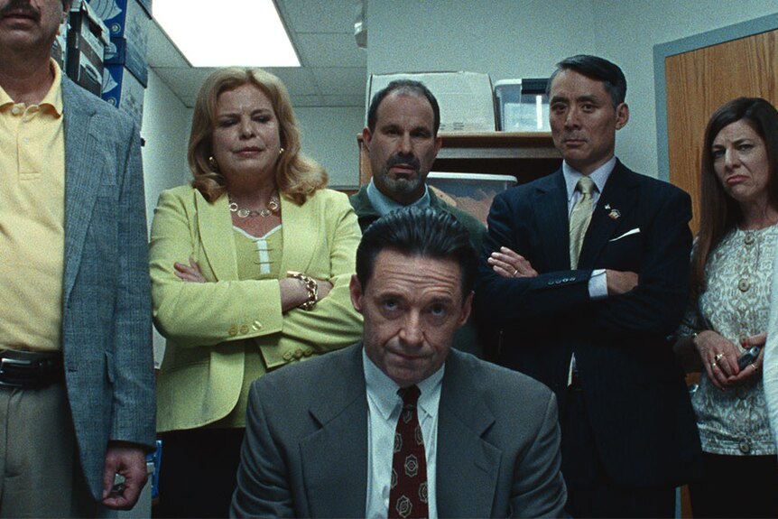 Small room with Hugh Jackman seated and behind him standing a line of six men and women looking unhappy, some with arms folded.