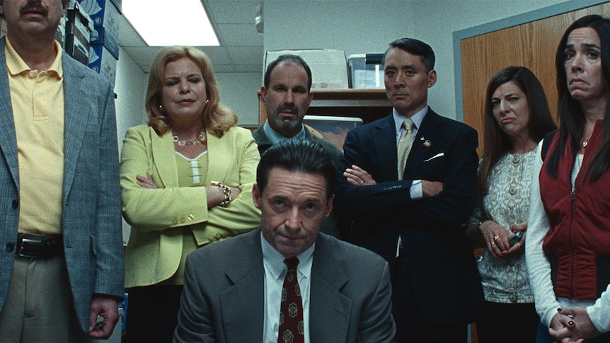 Small room with Hugh Jackman seated and behind him standing a line of six men and women looking unhappy, some with arms folded.