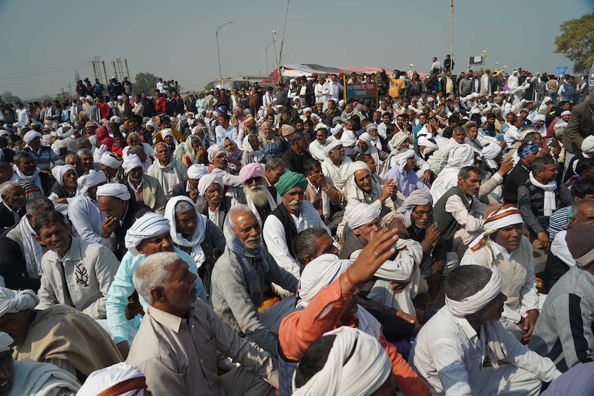 A large crowd of men, many wearing turbans, sit together on a road in protest.