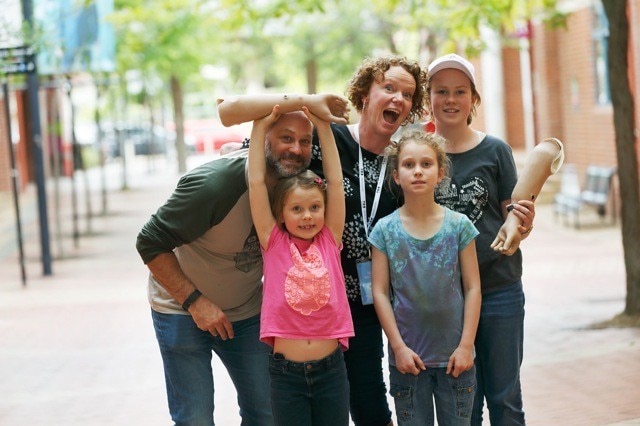 Two adults and three children huddle and act silly for the camera, two of the children hold prosthetic arms. Trees in background