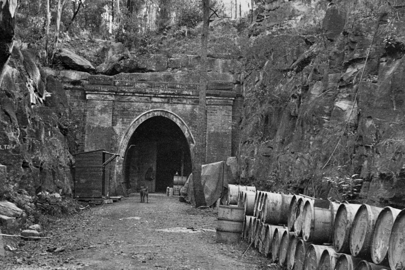 The entrance to a large underground tunnel, surrounded by rock walls, with storage drums in the foreground.