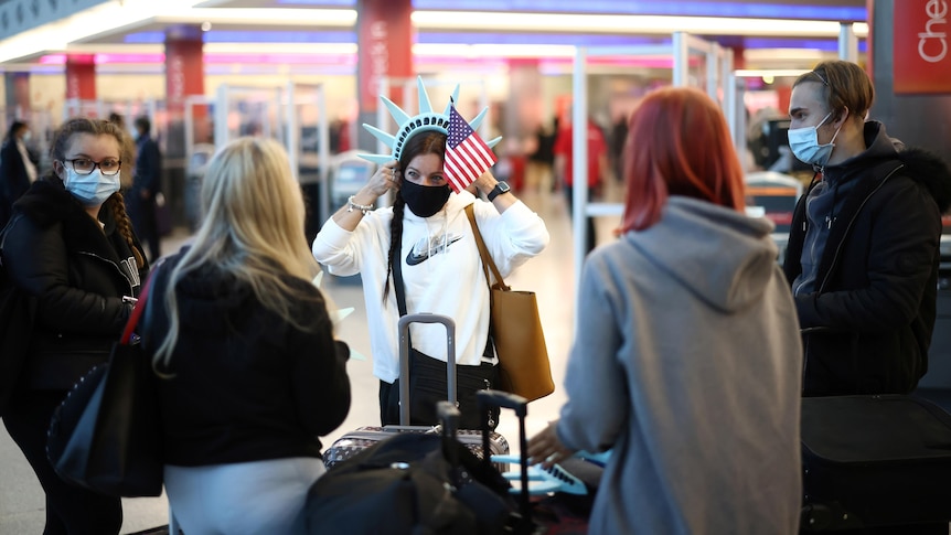 A group of people wearing face masks stand together in an airport near checkin desks. One wears a statue of liberty headband