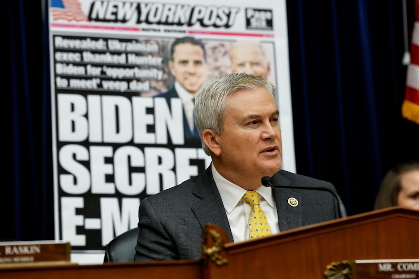 James Comer sits in front of a microphone. Behind him is a large image of a New York Post front page of Hunter and Joe Biden