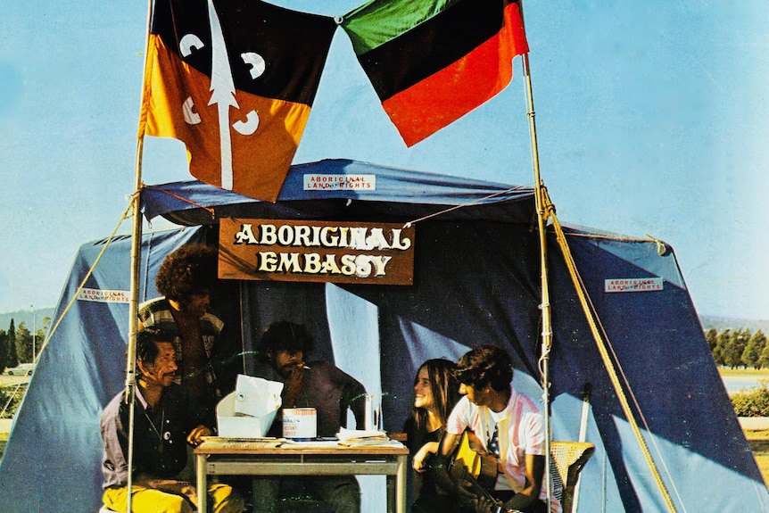 Five people are seen sitting at a table next to a blue tent with a sign on it reading "Aboriginal tent embassy" under two flags.