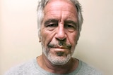 A middle-aged man with grey-white hair wearing a grey T-shirt looks serious in front of a plain background.