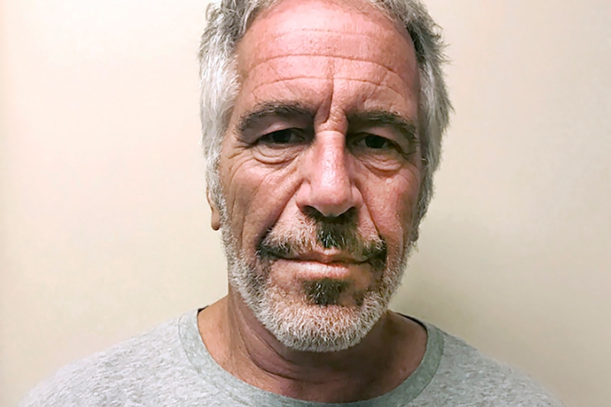 A middle-aged man with grey-white hair wearing a grey T-shirt looks serious in front of a plain background.