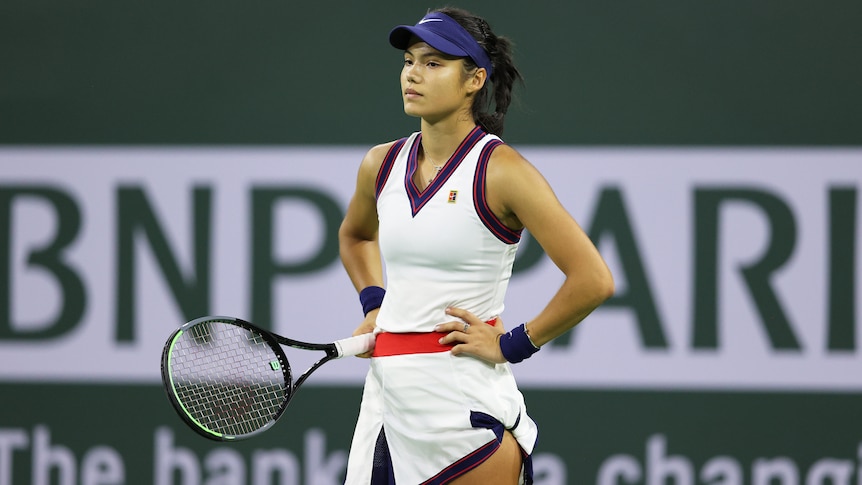 A female tennis player stands with her racquet in her right hand, with her hands on her hips.