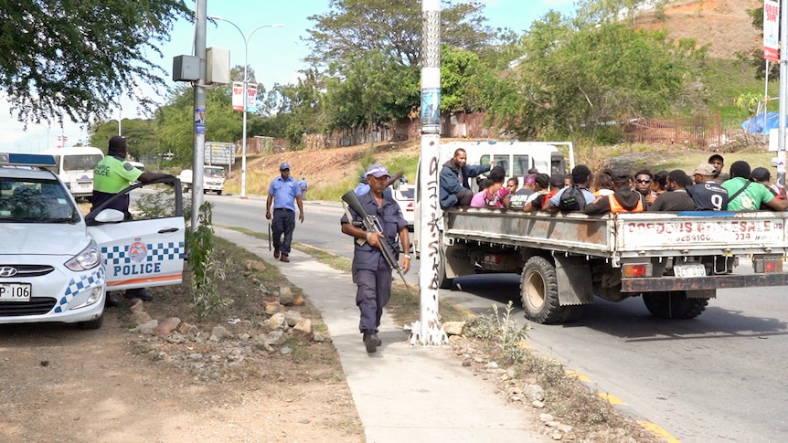 A group of people sit on the back of a truck as a man in uniform holding a gun walks by