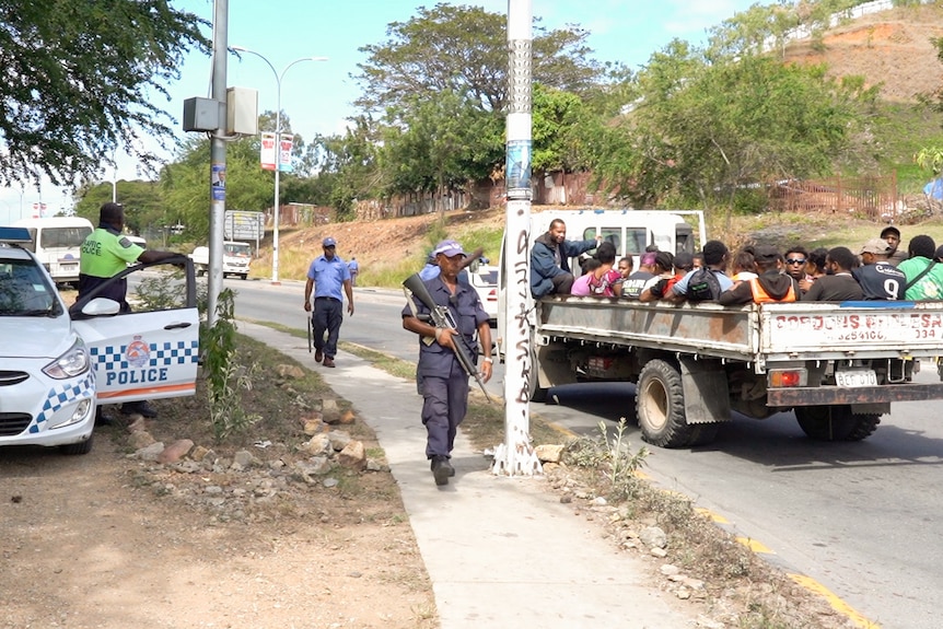 A group of people sit on the back of a truck as a man in uniform holding a gun walks by