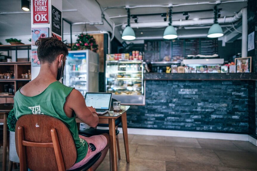 Man in singlet sits at computer in coffee shop.