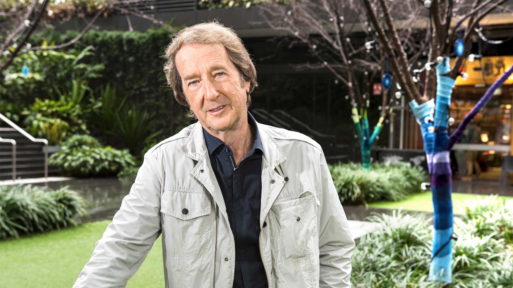 A man with blonde and brown hair, wearing a navy shirt and taupe jacket, standing in a city garden.