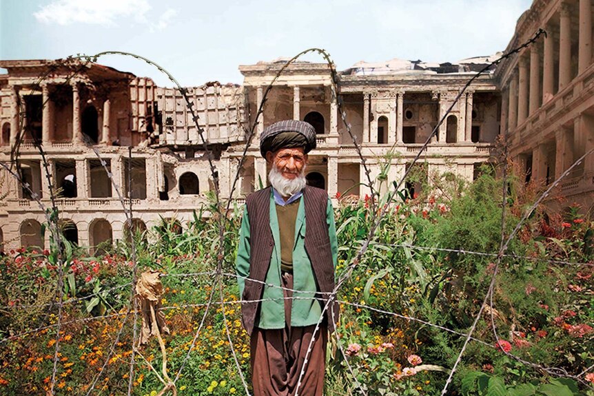 An Afghan man stands behind barded wire in a garden, with a ruined palace in the background.