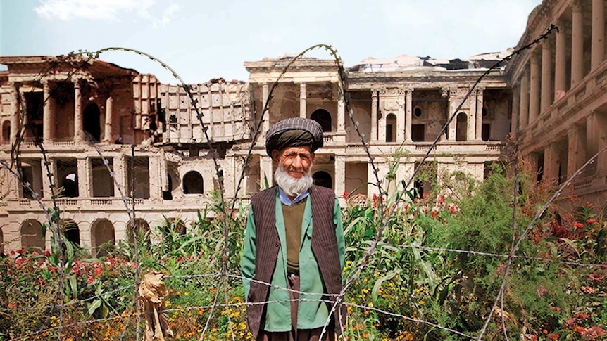 An Afghan man stands behind barded wire in a garden, with a ruined palace in the background.