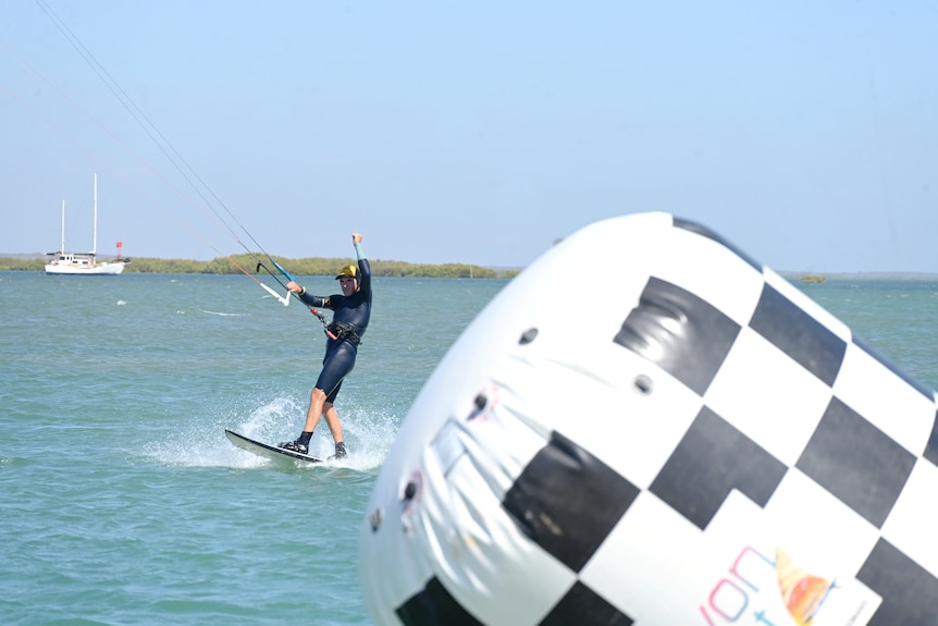 A man on a kitesurfing board speeds past a white and black checkered buoy raising a fist in celebration.