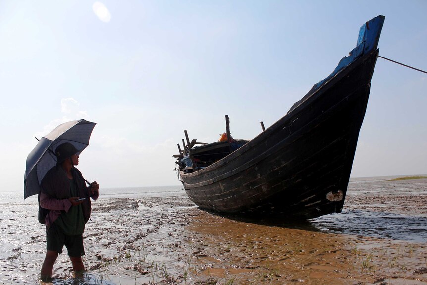A woman with an umbrella looks at a boat used to carry Rohingya refugees lies stranded in mudflats outside Yangon, Myanmar.