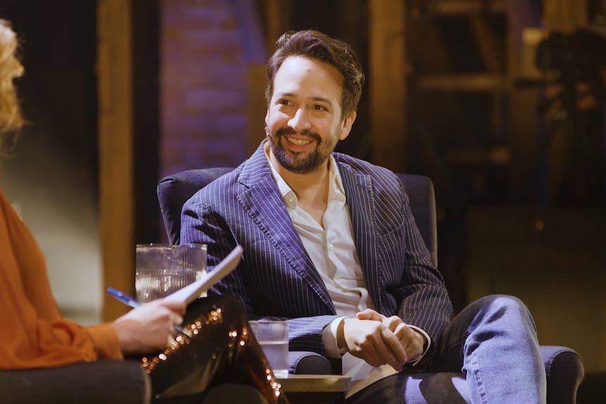 Lin-Manuel Miranda, a man in a blue striped jacket and white shirt, smiles whilst talking on stage to a woman in an orange shirt